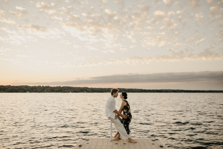 Engagement photographer takes picture of couple staring into each others eyes on a dock at the lake at sunset.