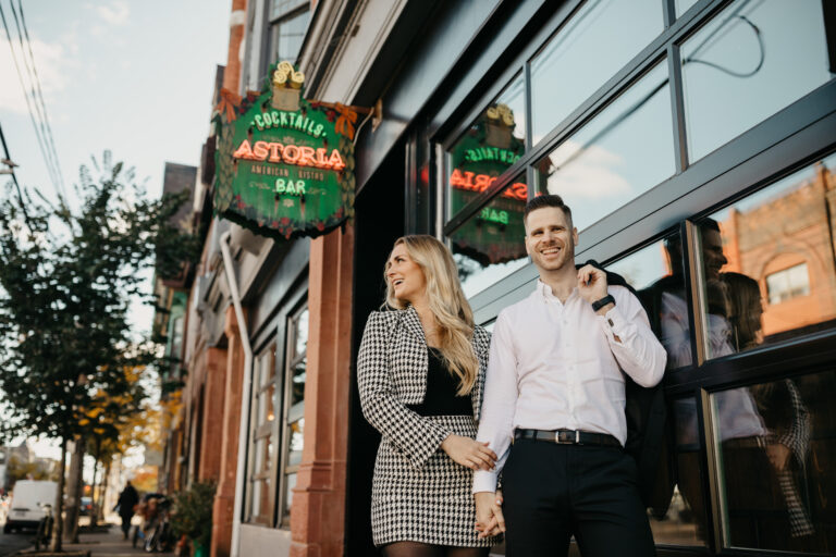 Couple walks down the street past a bar in Toronto Canada while engagement photographer takes a picture.